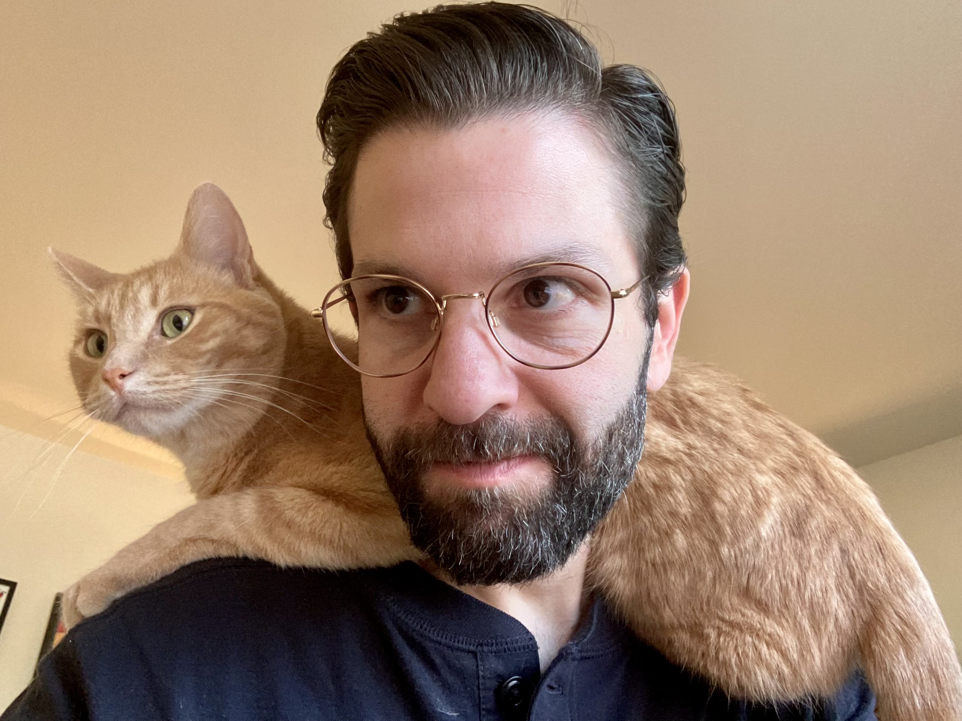 A portrait of me, with Teddy, an orange tabby cat, sitting across my shoulders. I'm wearing a dark blue shirt and gold-rimmed glasses.
