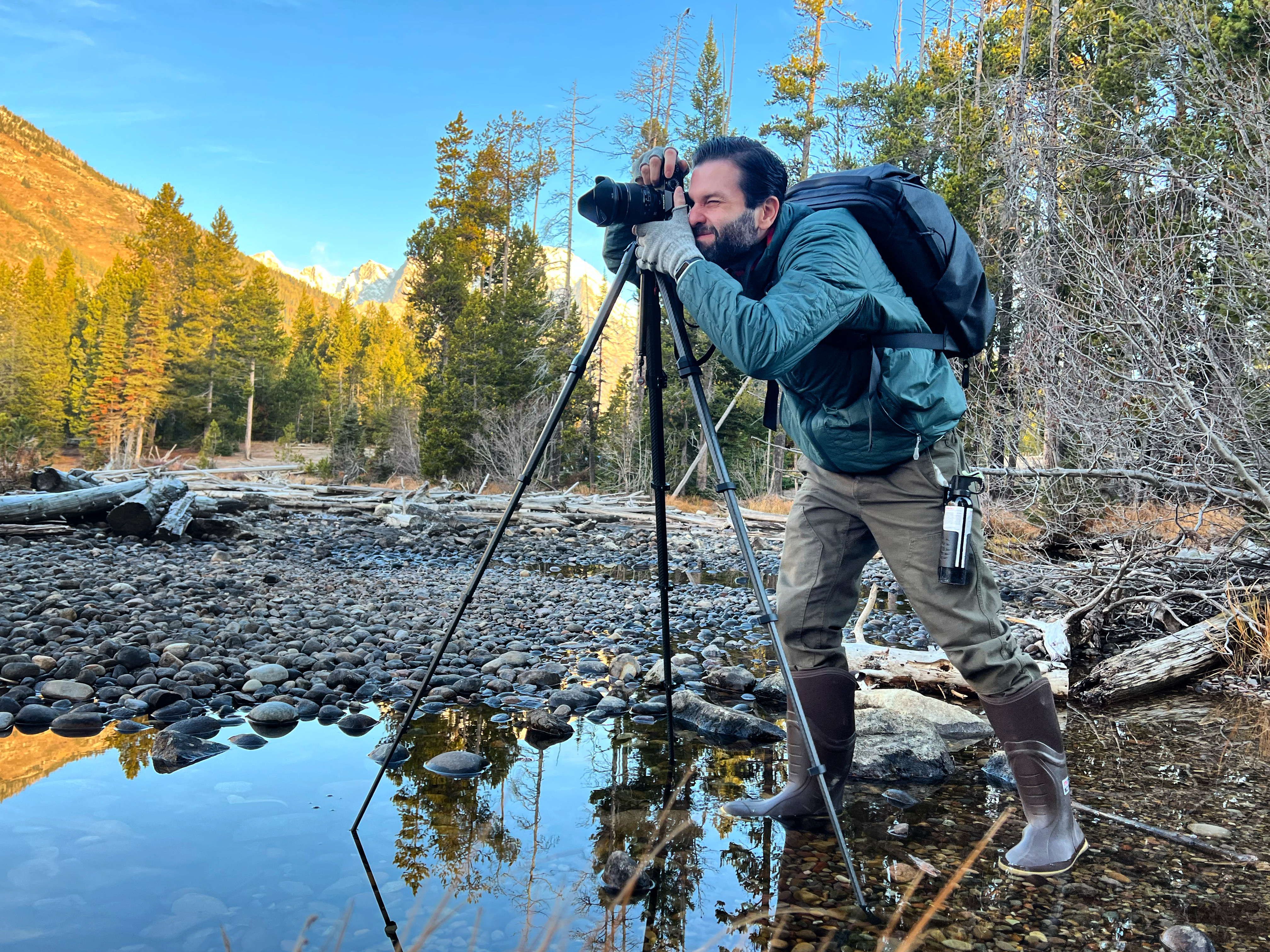 Me, looking through the viewfinder of my camera on a tripod, standing in water on a stream . In the background, a line of lodgepole pine trees, and behind them, Mount Moran. I'm wearing a green jacket and pants, green Xtratuf boots, gray gloves, a black backpack, and a can of bear spray.