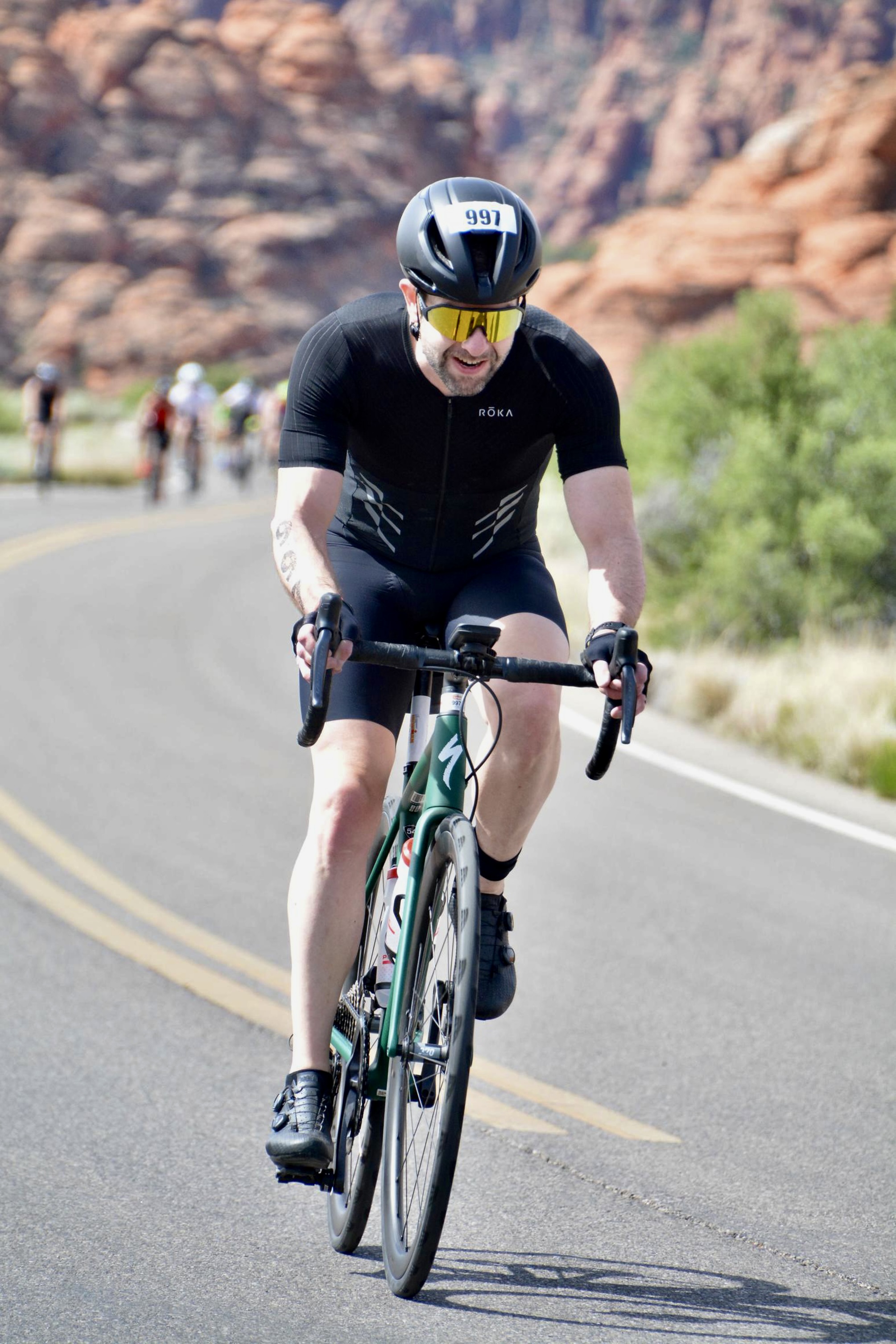 Front view of me climbing on a green Specialized Aethos bike through Snow Canyon. A group of cyclists can be seen in the background behind me. I'm wearing a black helmet, black trisuit, black gloves, black shoes, and gold sunglasses. I have a race tattoo in my right arm with the number 997. The helmet has a sticker on it with the same number.