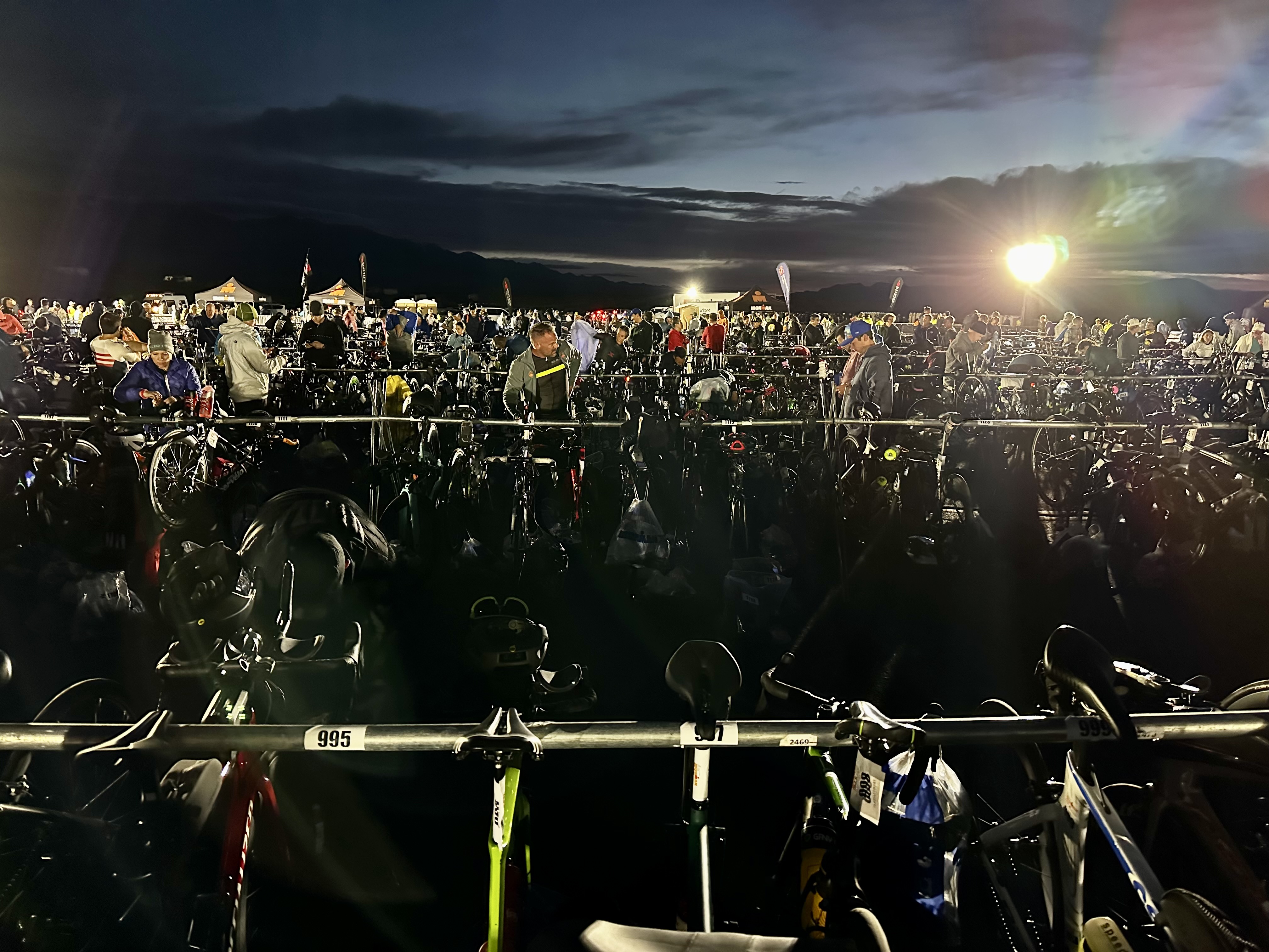 The T1 transition area for Ironman 70.3 St. George in Sand Hollow State Park, shortly before dawn. The racks with bikes can be seen, along with athletes setting up their gear before the swim.