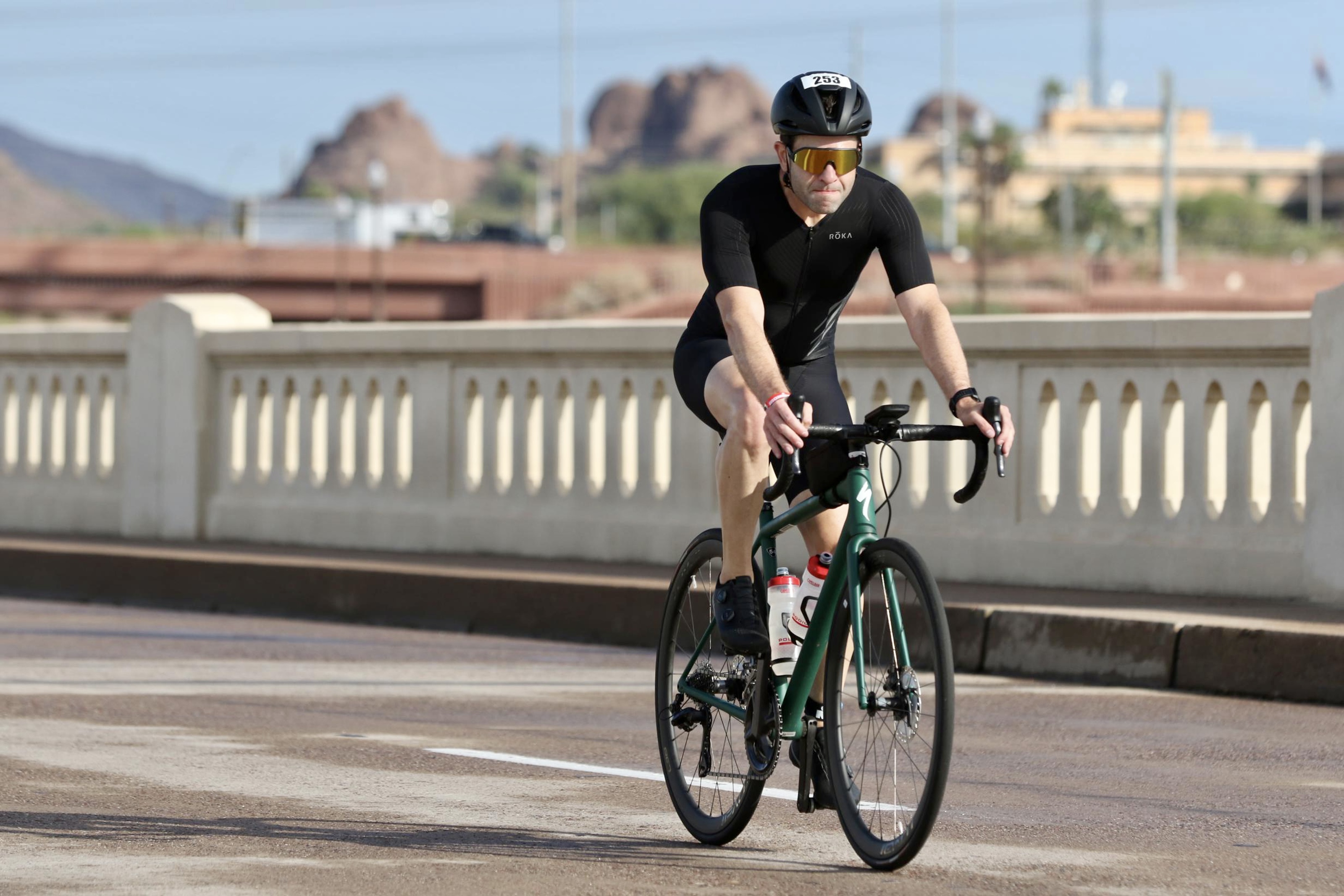 Me, riding a green Specialized Aethos bike on Mill Avenue bridge in Tempe. I'm wearing a black helmet, gold sunglasses, black tri suit, and black shoes.