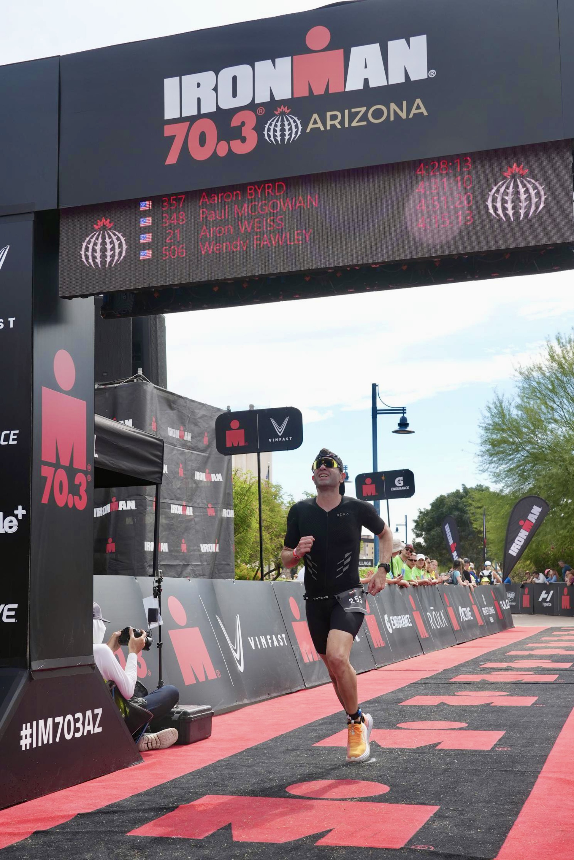 Me, about to cross the finish line at Ironman 70.3 Arizona, running on a red carpet with the M-dot logo and under the finish line arch, with a pained expression on my face.