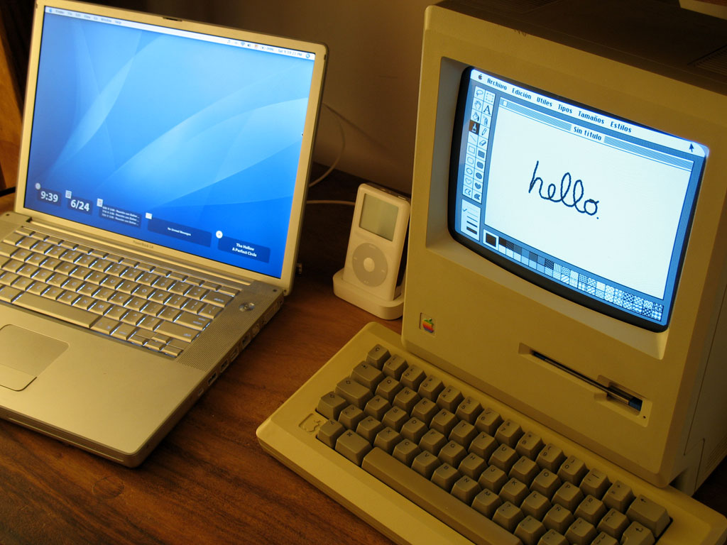 A Macintosh 512K next to a G4 PowerBook on a wooden desk. A fourth-generation iPod sits on a cradle between them.