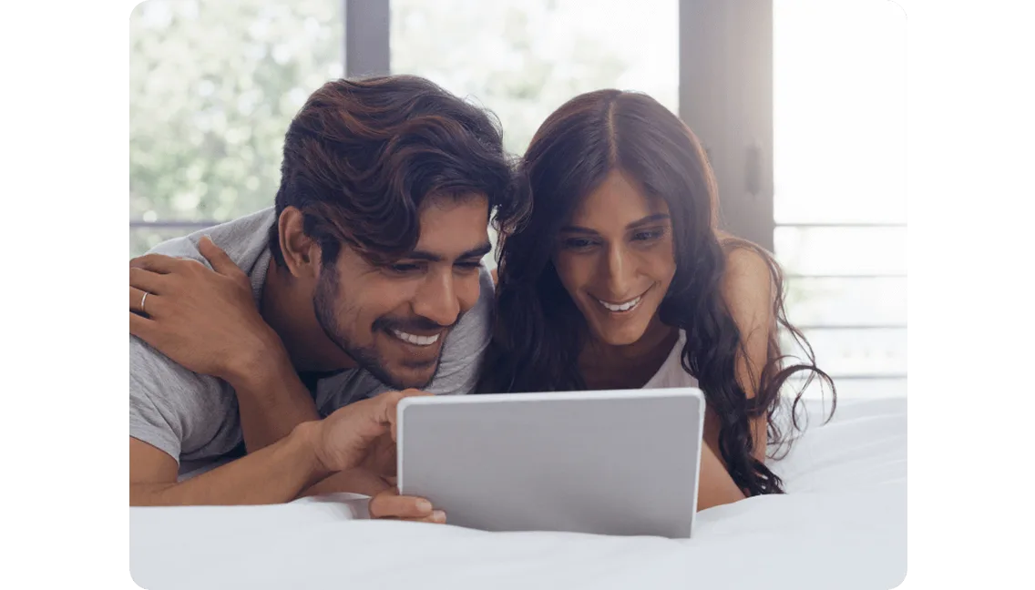 Two people lie on a bed, smiling as they watch something on a tablet.