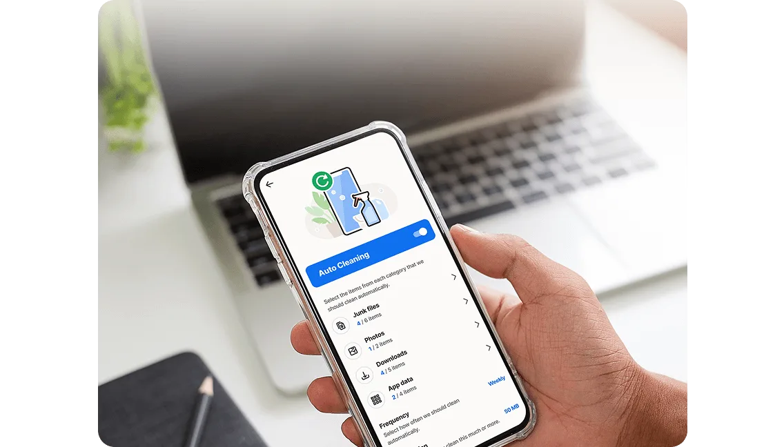 A hand holding a smartphone displaying the Norton Utilities Ultimate app with the "Auto Cleaning" feature enabled. A laptop is blurred in the background.
