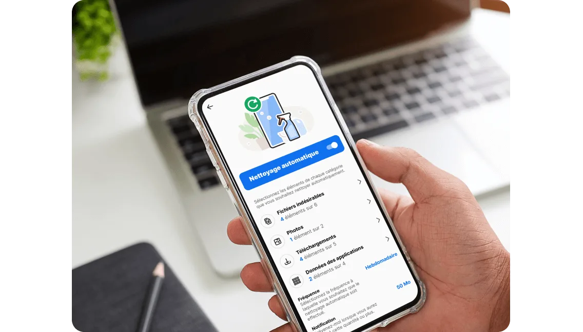 Une main tenant un téléphone intelligent qui affiche l’application Norton Utilities Ultimate avec la fonction de nettoyage automatique activée. Un ordinateur portable est flou en arrière-plan.