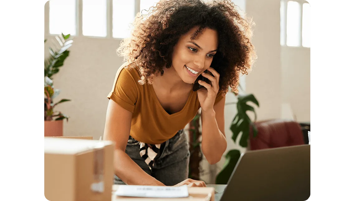 A person leaning over a desk while speaking on the phone and using a laptop. Cardboard boxes and plants are visible in the background.