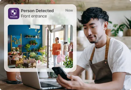 Shop owner checks phone; TELUS alert overlay reads “Person Detected — Front entrance.”