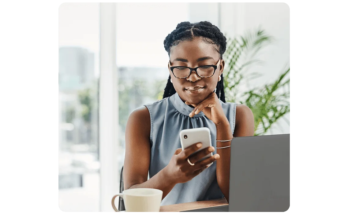 A person seated at a desk, holding a smartphone and using a laptop, with a coffee mug nearby. A bright office setting is in the background.