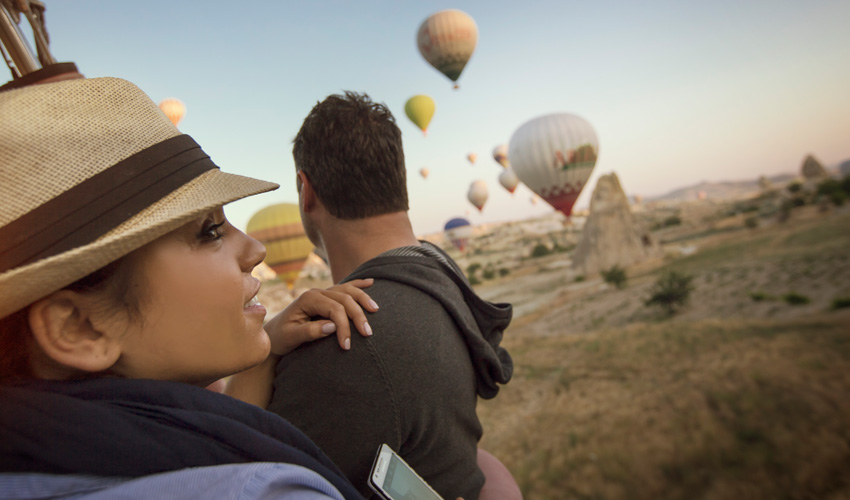 A couple riding a hot air balloon enjoys a scenic view of a balloon-filled sky.