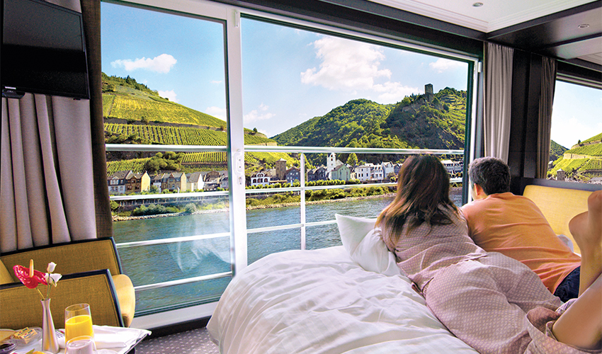 Avalon Waterways - A couple in pyjamas lie on their stateroom bed and admire the river views