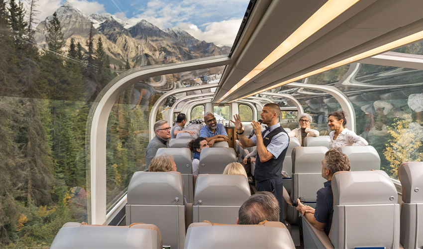 Rocky Mountaineer® - Tourists listen to a guide describe the scenery from the panorama train lounge