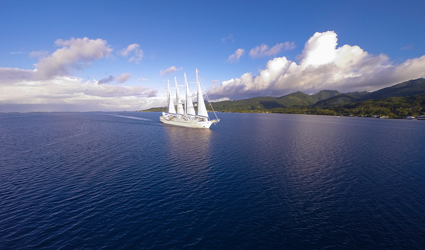 Windstar® - Aerial view of a sailing yacht with sails up surrounded by blue skies and green mountains