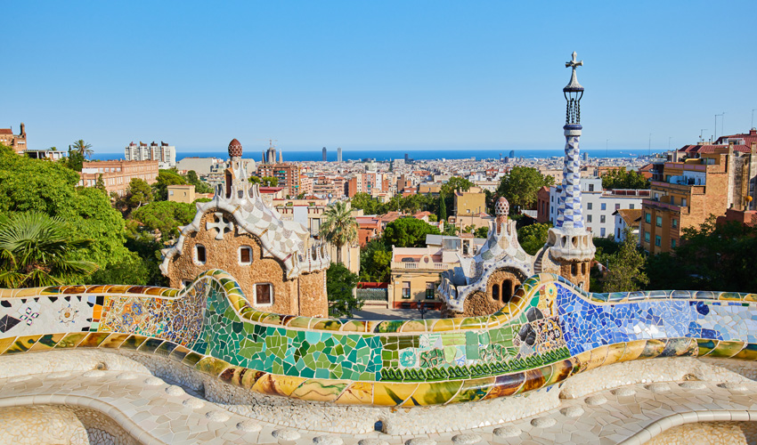 A view of mosaic benches at Park Guell in Barcelona with the city skyline and the sea in the background.