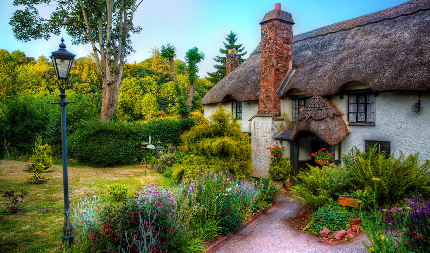 Exterior of a charming thatched roof cottage surrounded by lush English gardens.