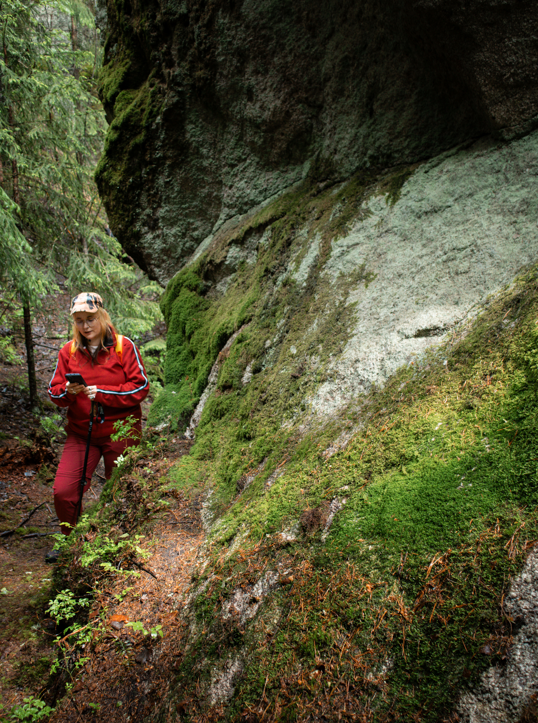 A person in a red hiking outfit stands next to a mossy rock and reads a poem from their phone