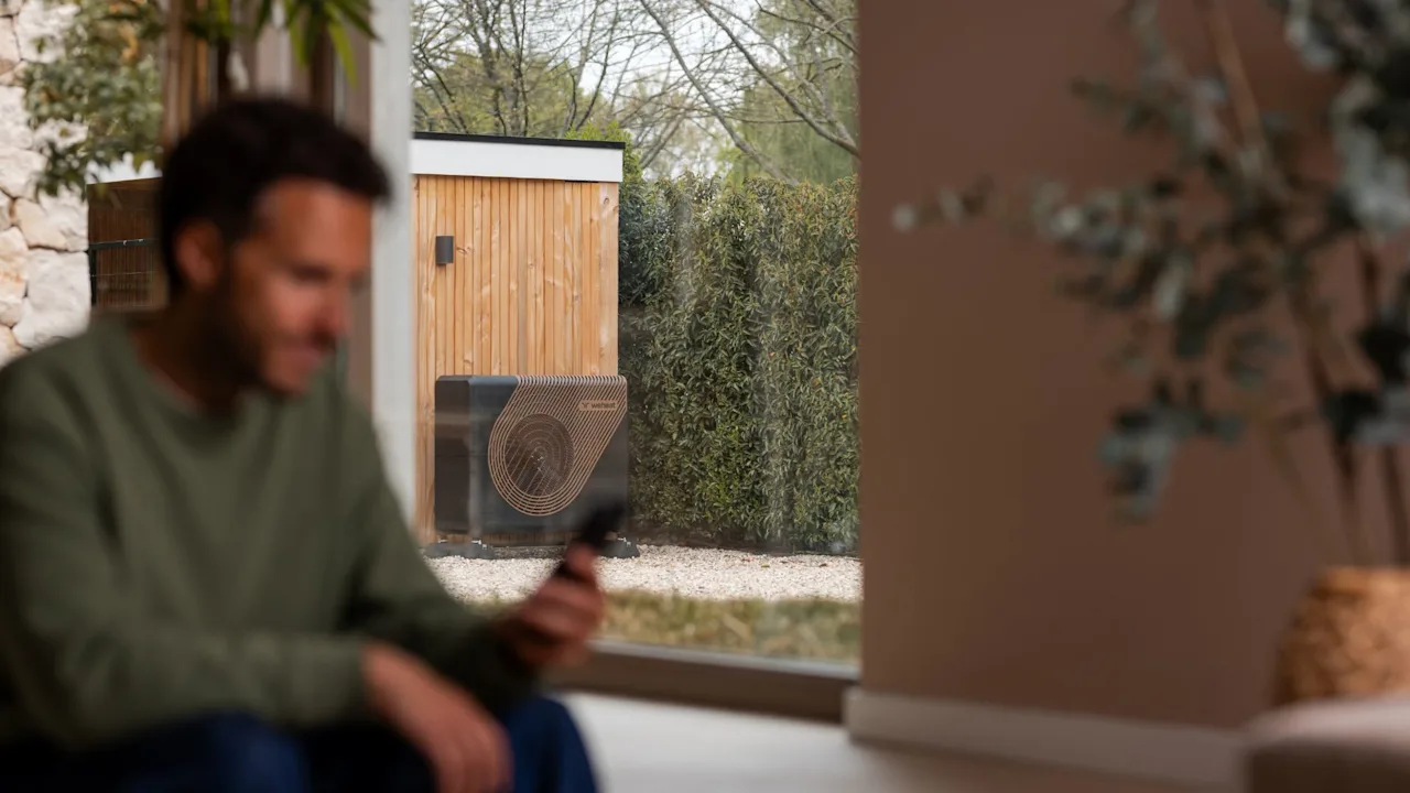 Afbeelding met een man die kijkt op de Weheat app op zijn telefoon met in de achtergrond die Sparrow warmtepomp in de tuin