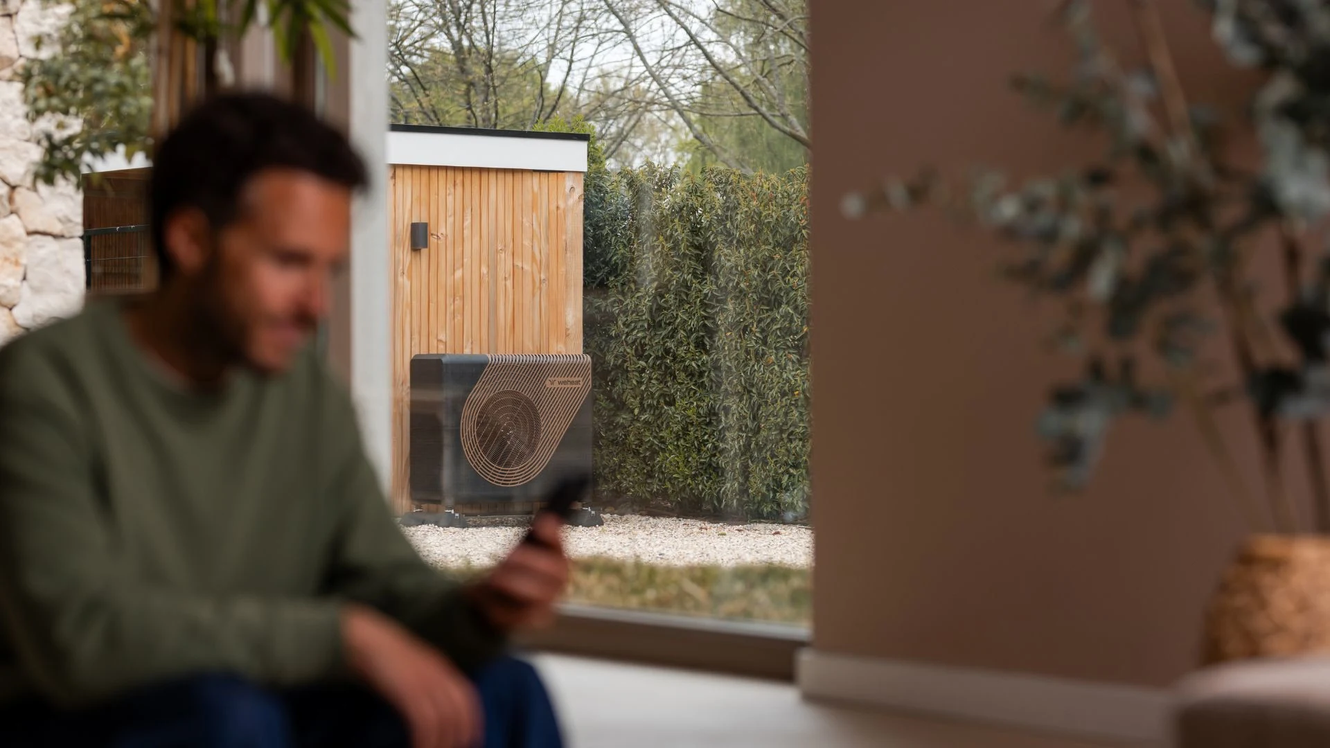 Afbeelding met een man die kijkt op de Weheat app op zijn telefoon met in de achtergrond die Sparrow warmtepomp in de tuin