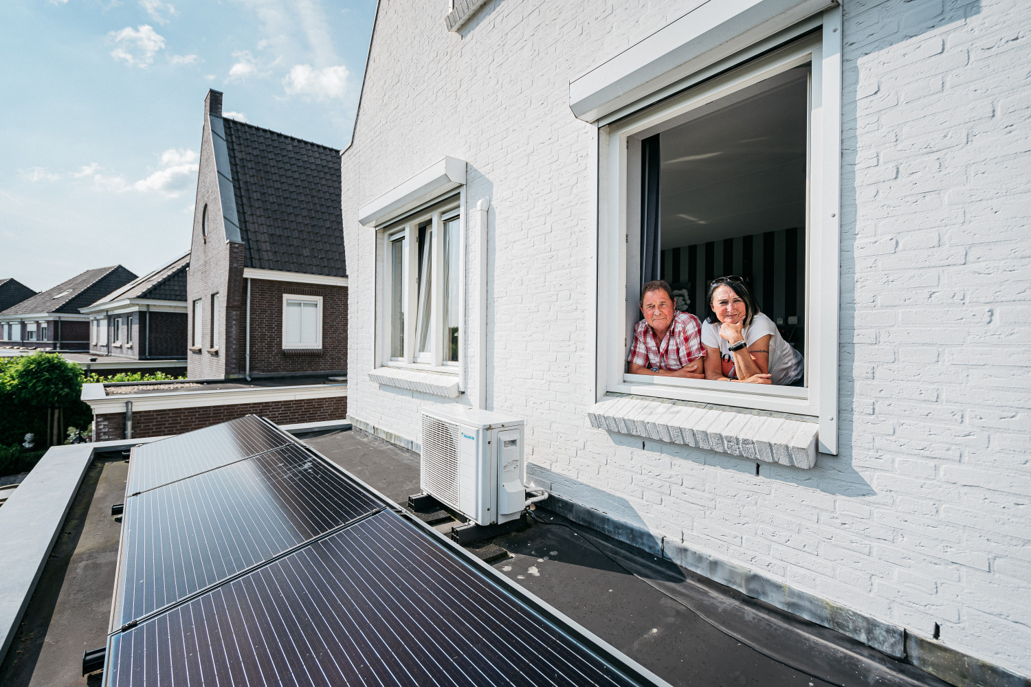 2 blije klanten kijken uit het raam over hun platte dak met zonnepanelen en een warmtepomp