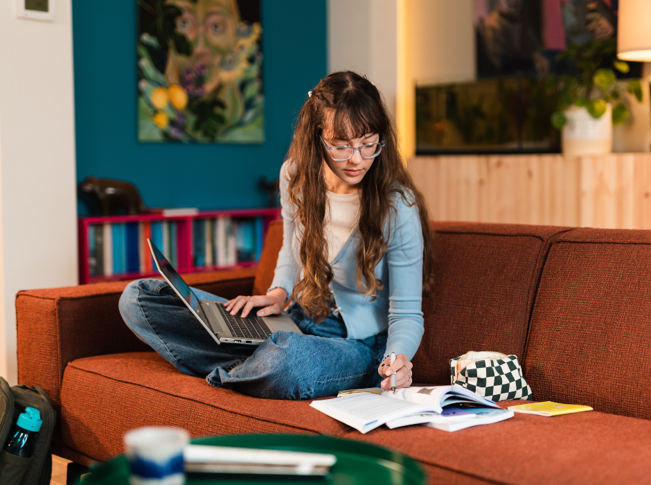 Meisje zit op de bank met haar schoolspullen en kijkt in haar boeken met haar laptop op schoot