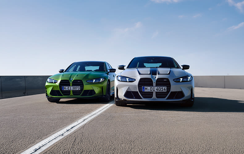 Front view of green BMW M3 Sedan and white BMW M4 Coupé parked side by side on a rooftop under clear skies.