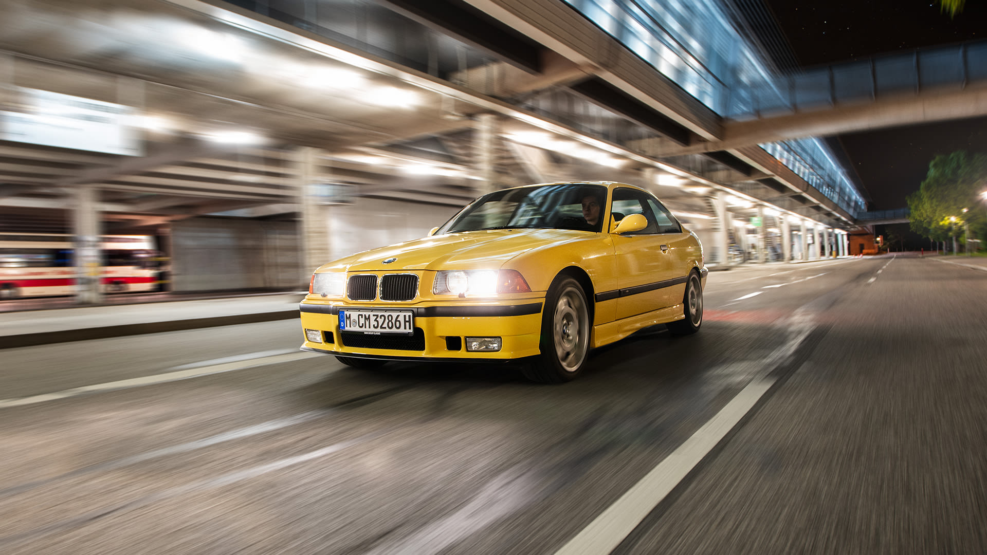 Yellow BMW Classic driving in the evening on a street with illuminated shops on the left.