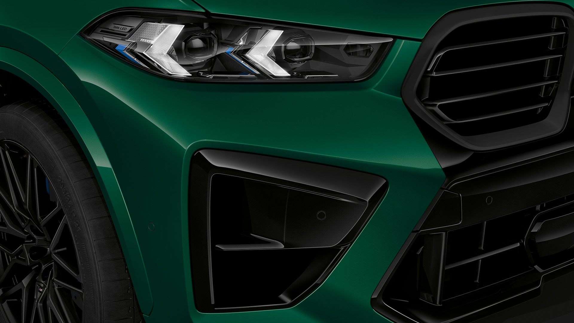 Close-up of the front right side of a green BMW SUV, showing the headlight, grille, and part of the front wheel.