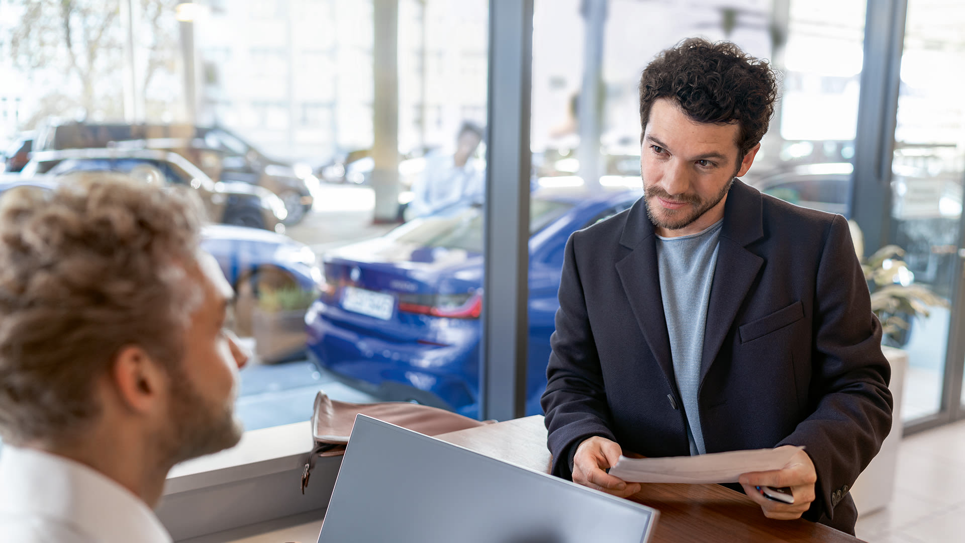 Person reviewing paperwork across a desk with another person in a bright BMW car showroom, with vehicles in the background.