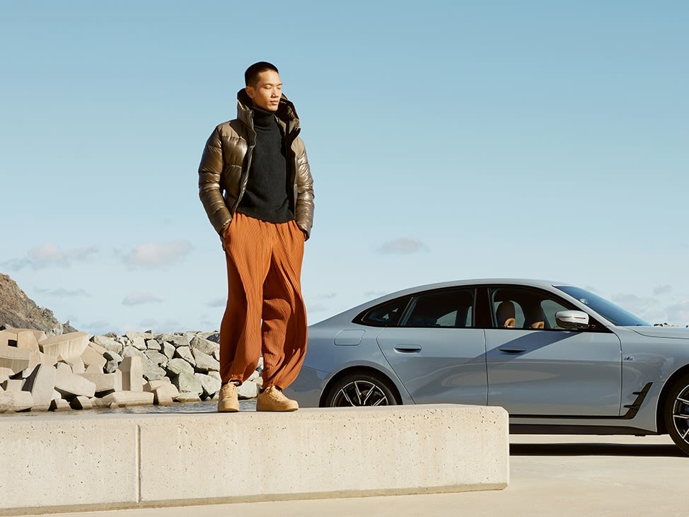 Person standing on a concrete platform wearing a puffer jacket and orange pants, with a silver BMW car parked nearby and a rocky landscape in the background.