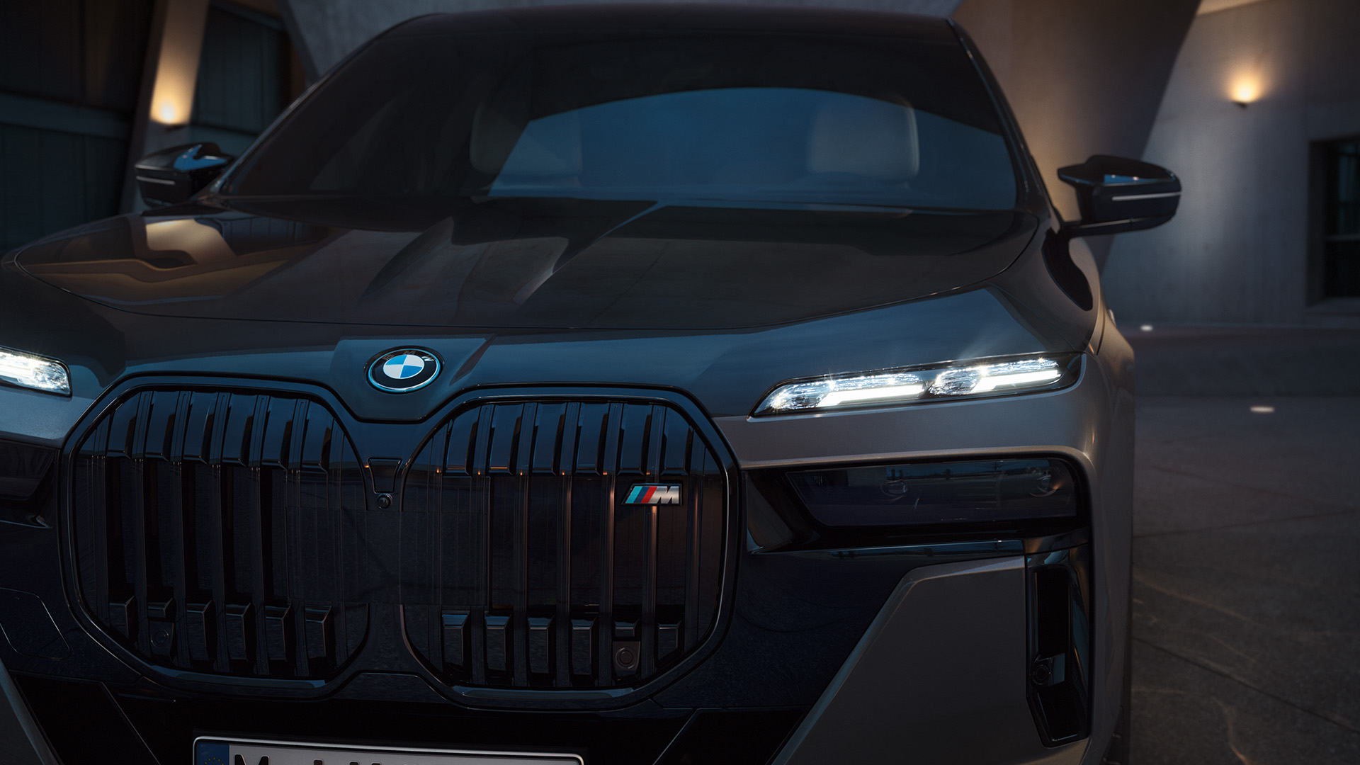 Front view of a black BMW car with illuminated headlights, parked in a modern architectural setting at dusk.