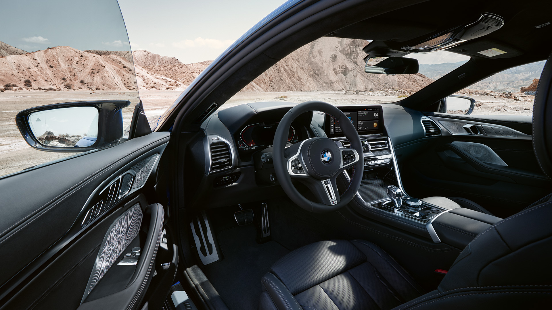 Interior view of a luxury BMW car with a sleek black leather interior, showing the steering wheel, dashboard, and center console. The car door is open, revealing a desert landscape in the background.
