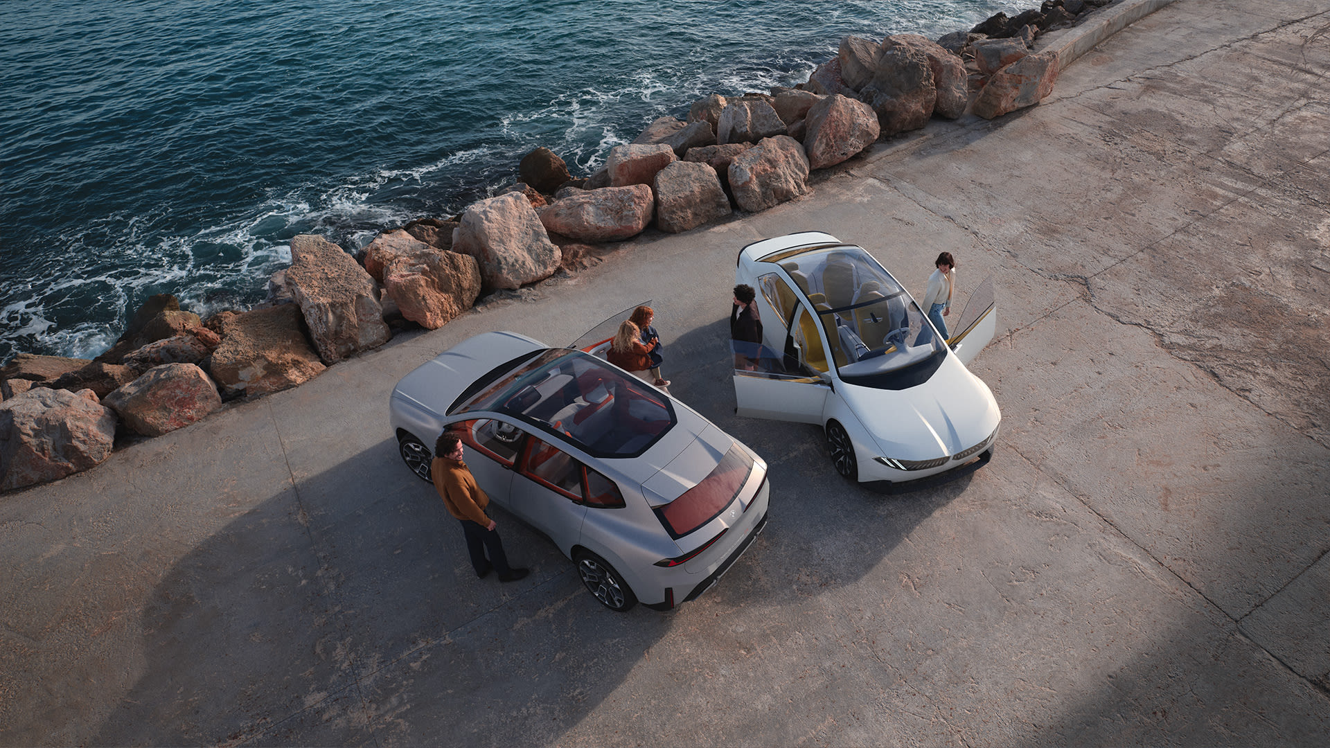 Silver and white BMW cars by the sea with people gathered around on a concrete surface
