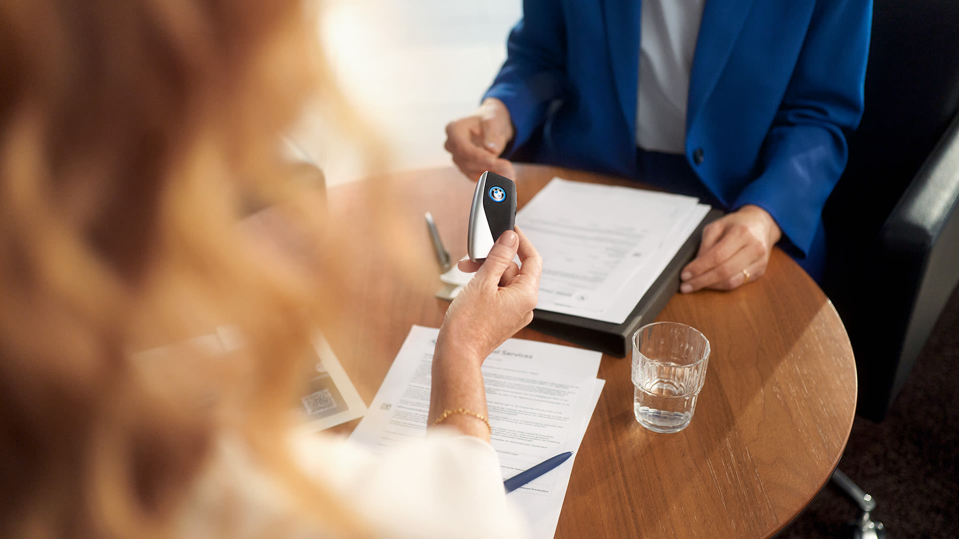 Two presons facing each other sitting at a round table with contracts on it and one person is handing a BMW key to the other.