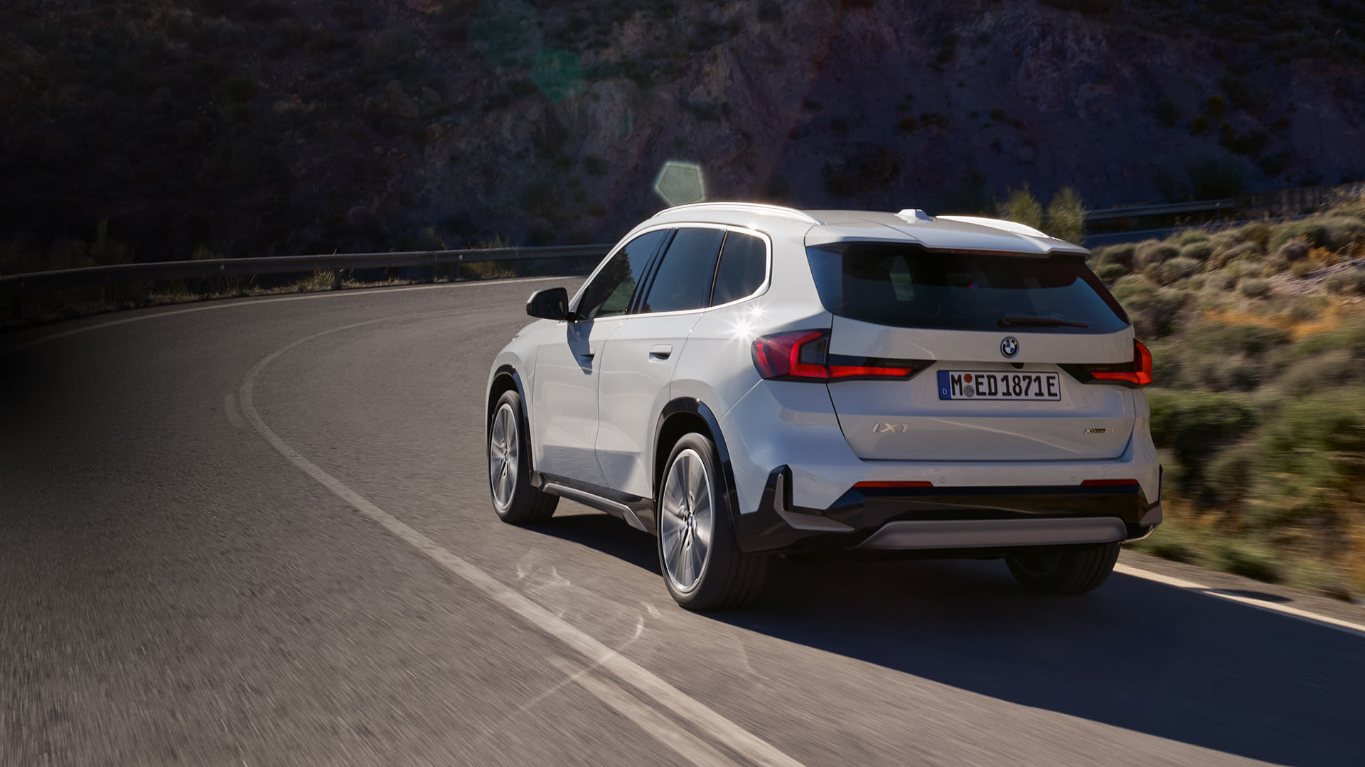 White BMW iX1 driving on a winding mountain road in a dry, rocky landscape, viewed from the rear with sunlight and motion blur conveying movement.