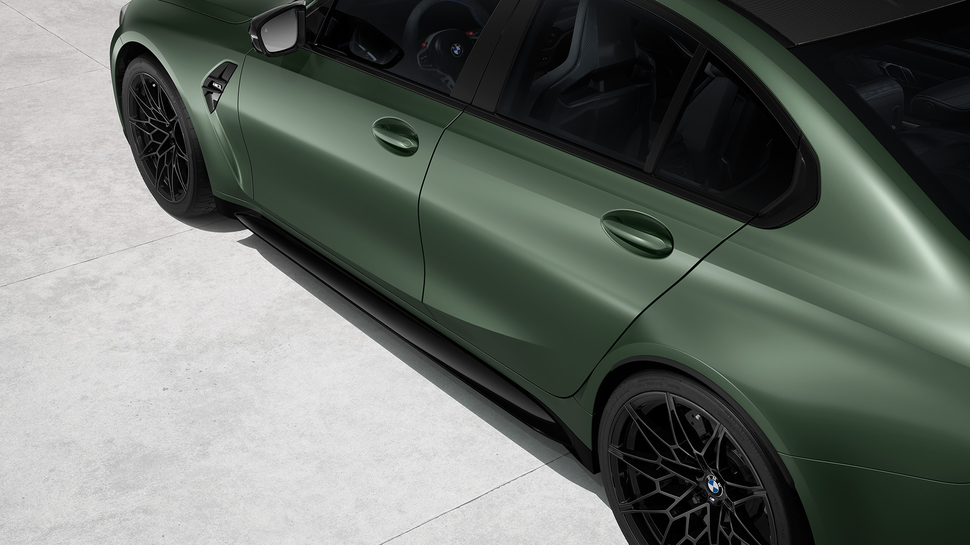 Top view of a matte green BMW saloon with black rims and a carbon fiber roof, parked on a concrete surface.