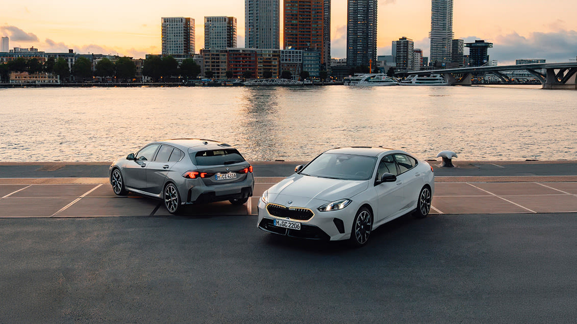 A white BMW 2 Gran Coupe car and grey BMW 1 Series car parked beside a riverside road overlooking water and a modern city bridge at sunset.