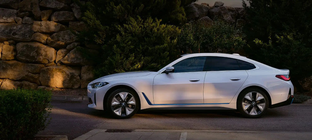 White modern BMW Saloon parked roadside, side profile view with stone wall and greenery backdrop.