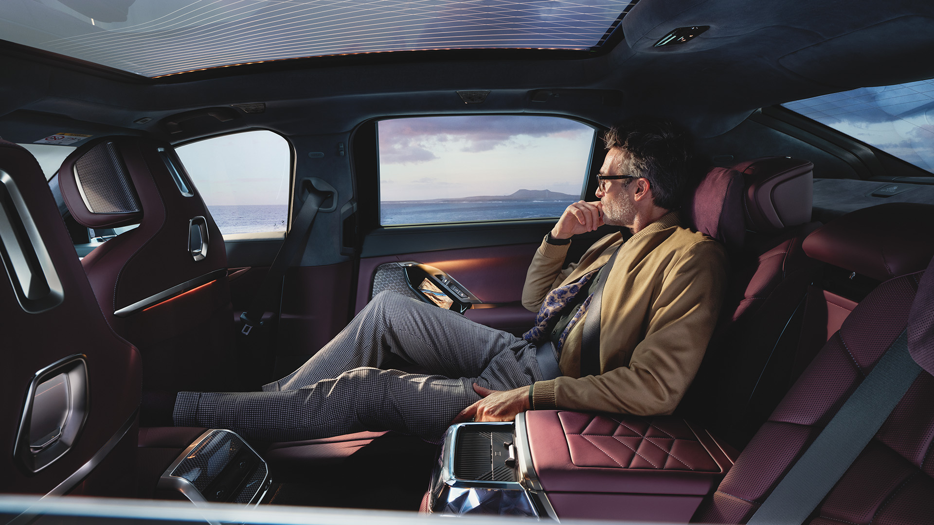Interior of a luxury car with a person reclining in the back seat, featuring plush maroon upholstery, ambient lighting on the ceiling, and large windows showcasing an ocean view.