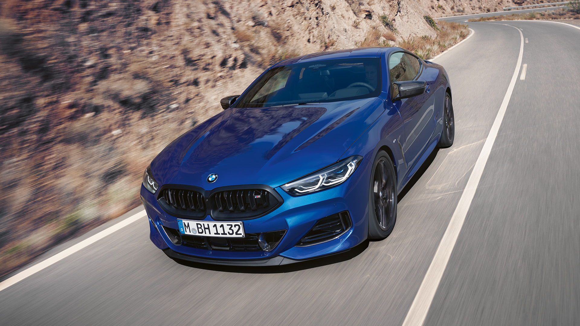 A blue BMW 8 series coupé driving on a winding road through a mountainous desert landscape.