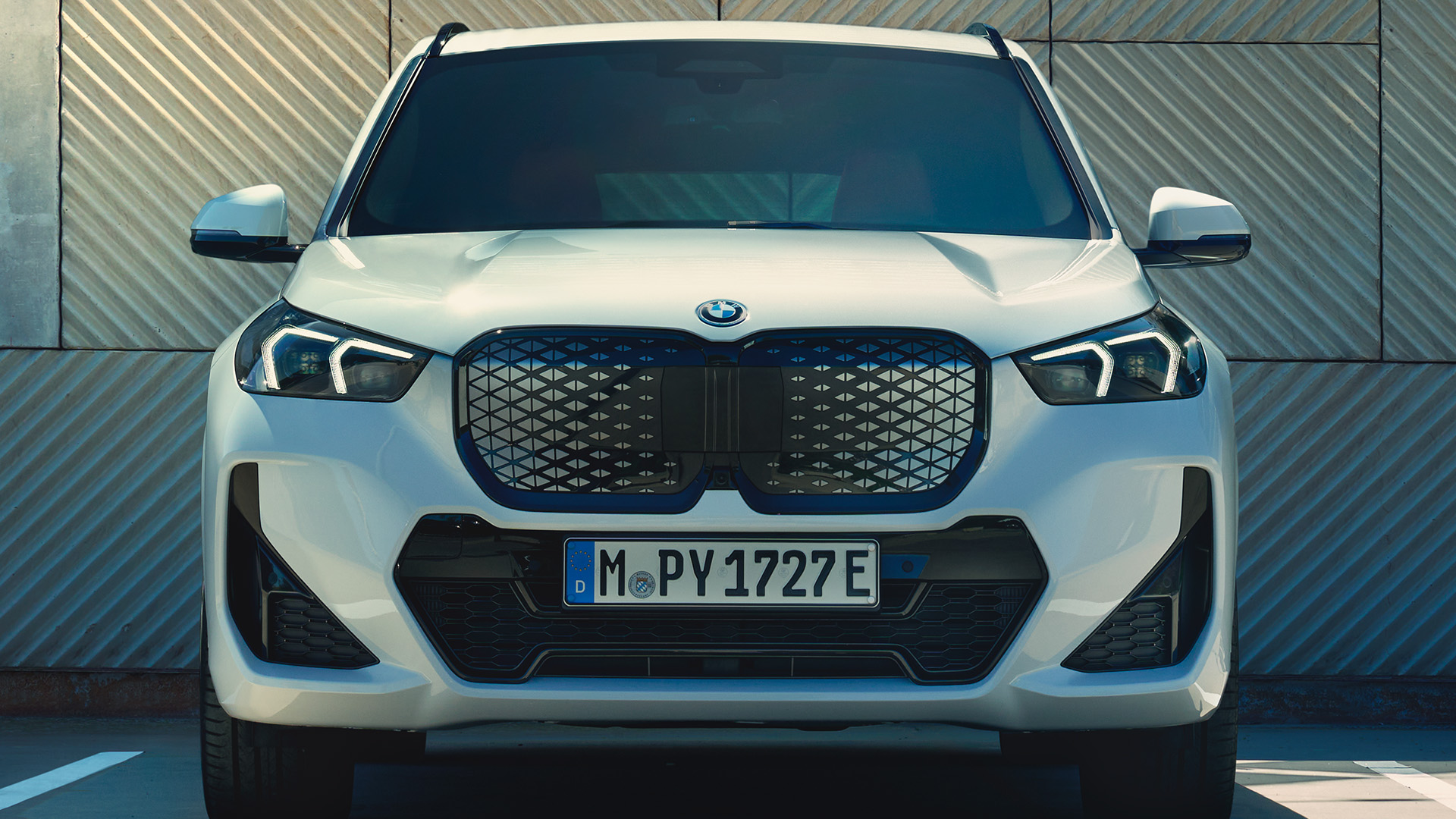 Front view of a white BMW car parked in an empty parking space with a modern, geometric-patterned building in the background.