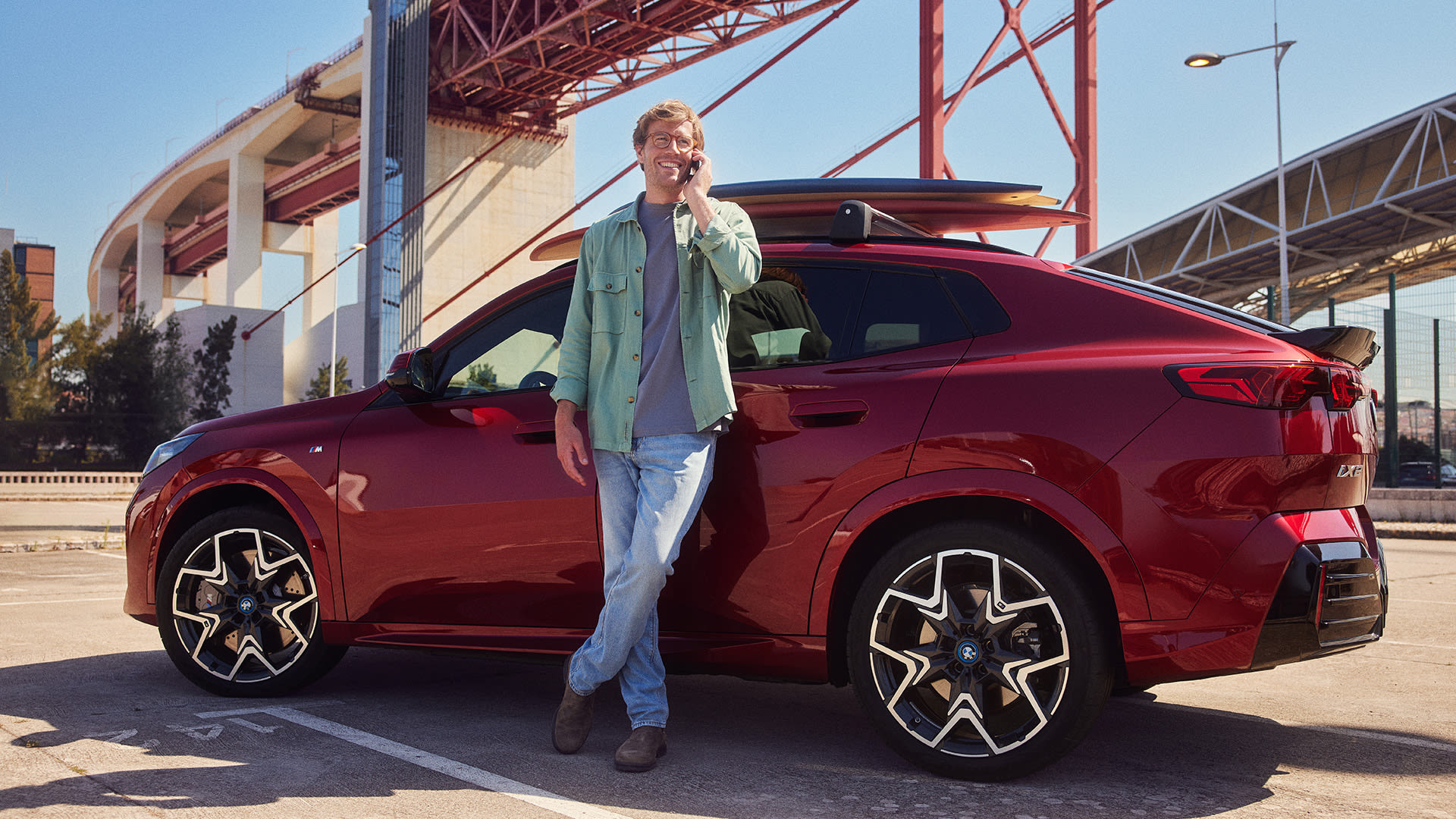 Person leaning against a red BMW SUV while using a mobile phone in an urban setting with industrial architecture.