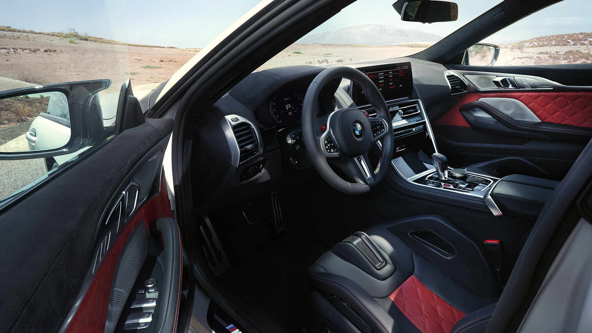 Interior view of a BMW M8 Competition Gran Coupé car, showcasing the steering wheel, dashboard, and center console with red and black leather seats and door panels. The car is parked in a desert landscape with mountains in the background.