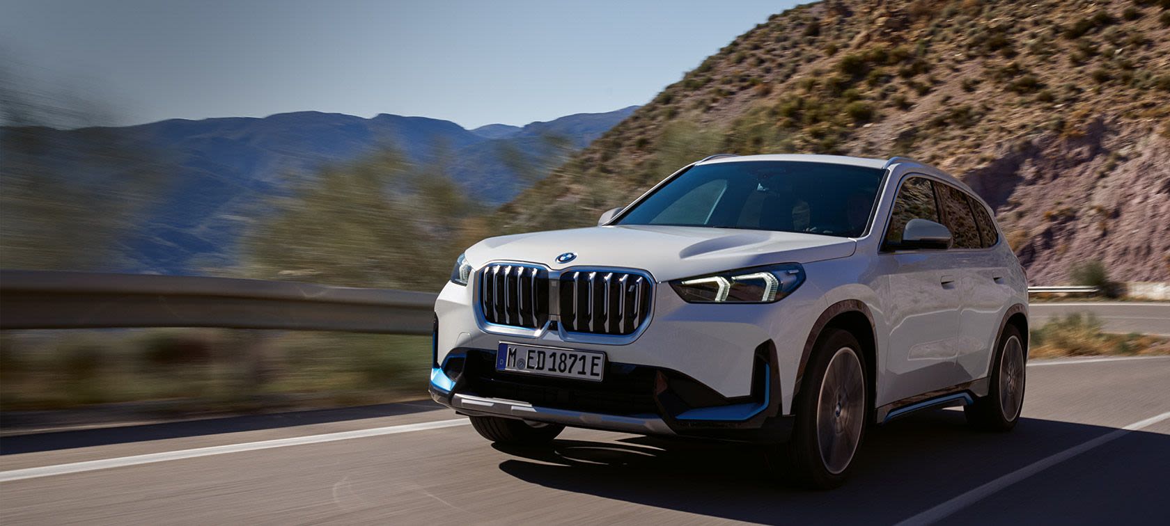 A white BMW SUV driving on a mountain road with a scenic background of hills and clear blue sky.