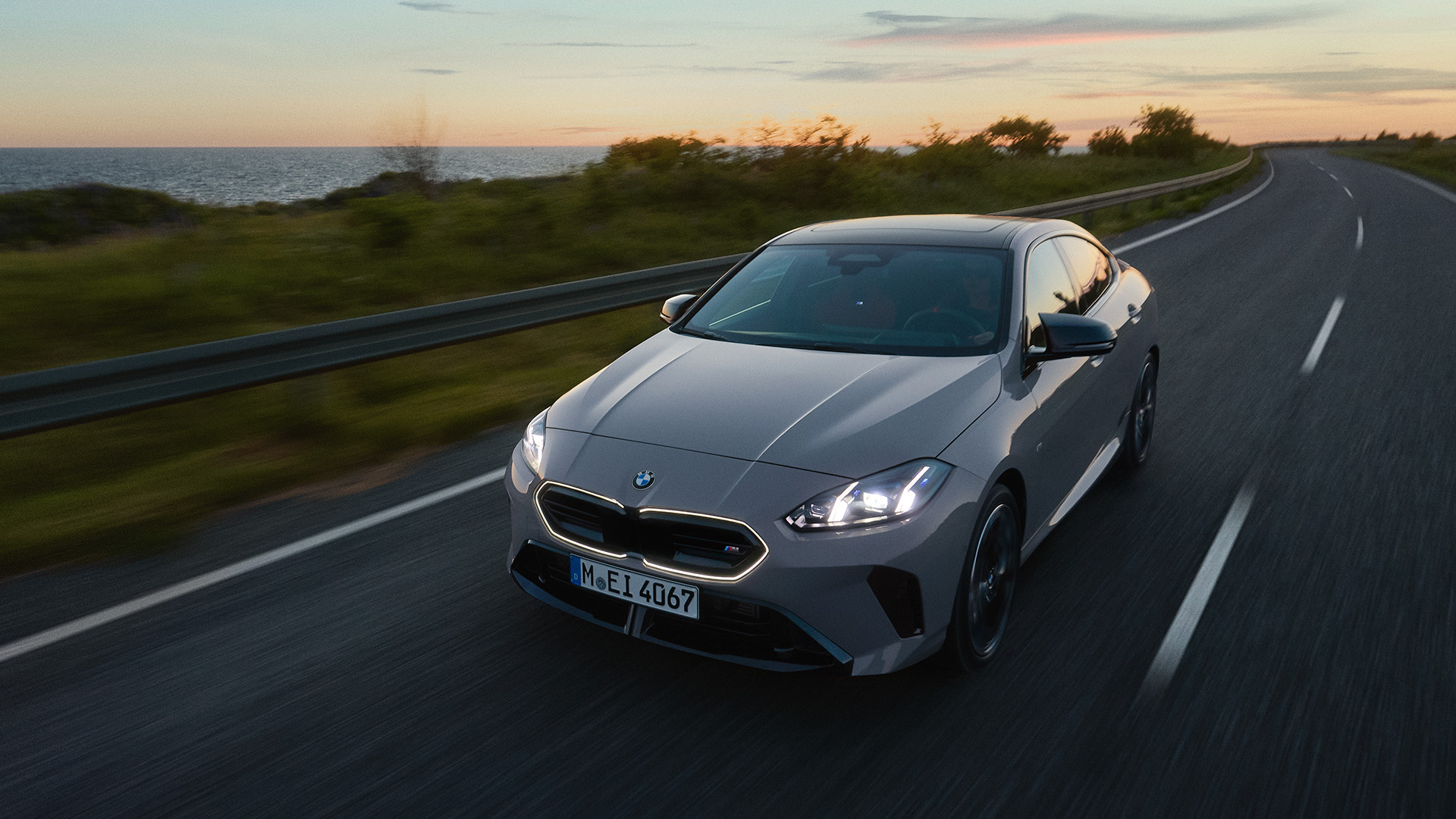 A silver BMW car driving on a scenic coastal road at sunset with the ocean and greenery in the background.