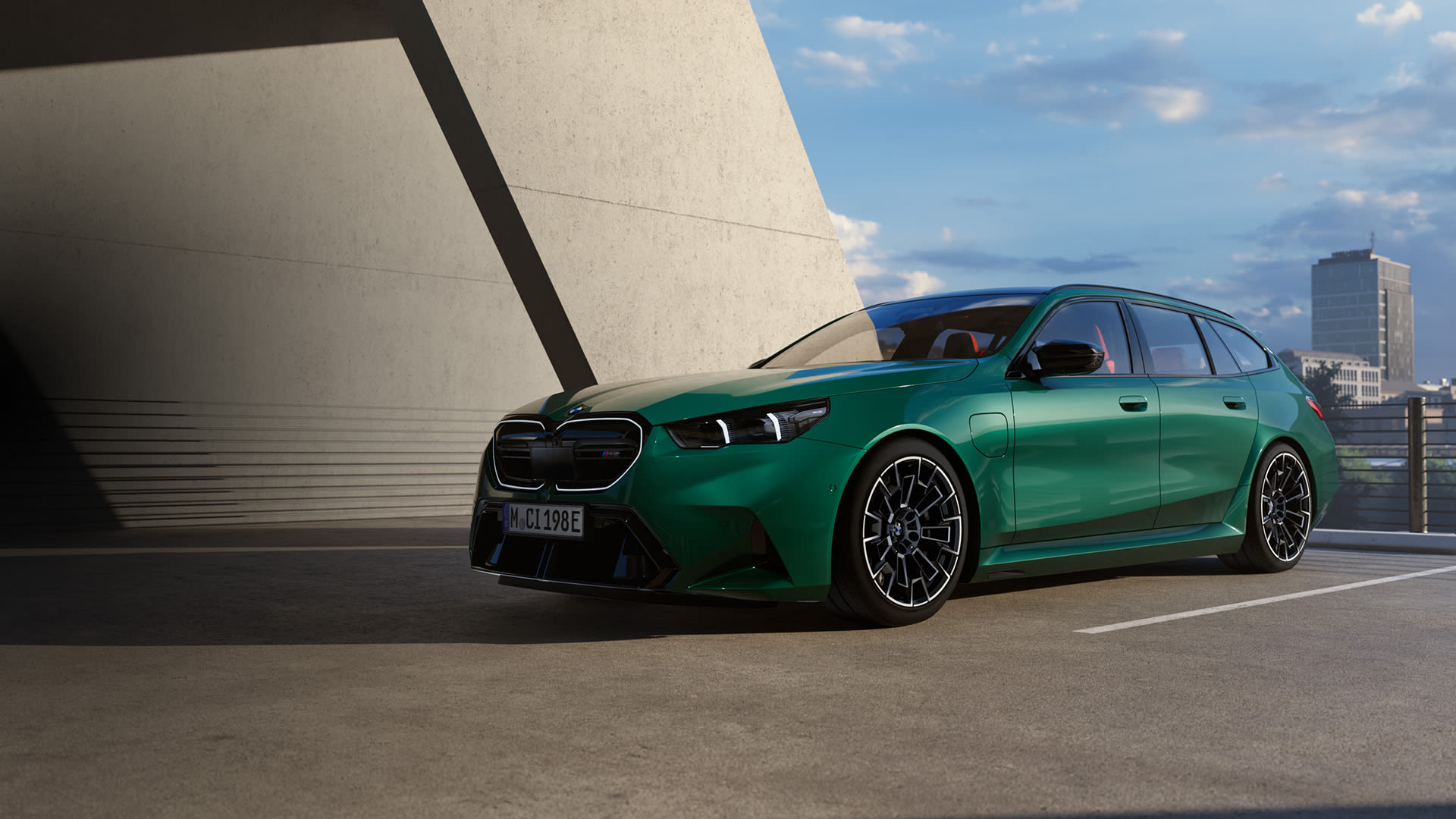 Green BMW M5 Touring car parked in a modern concrete rooftop car park, with city buildings visible in the background and sunlight casting shadows.