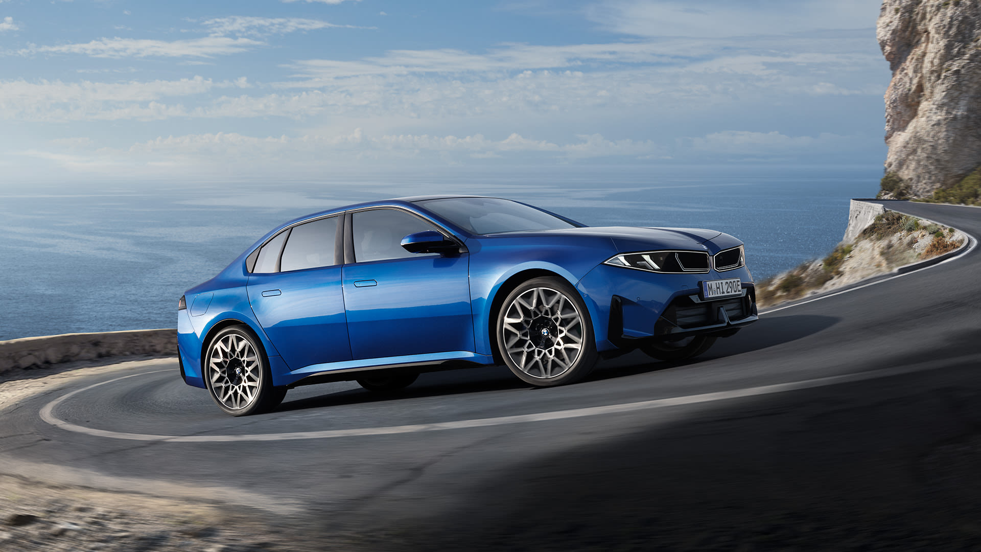 Blue BMW i5 saloon driving on a winding coastal road, with sea views and rocky cliffs under a bright sky.
