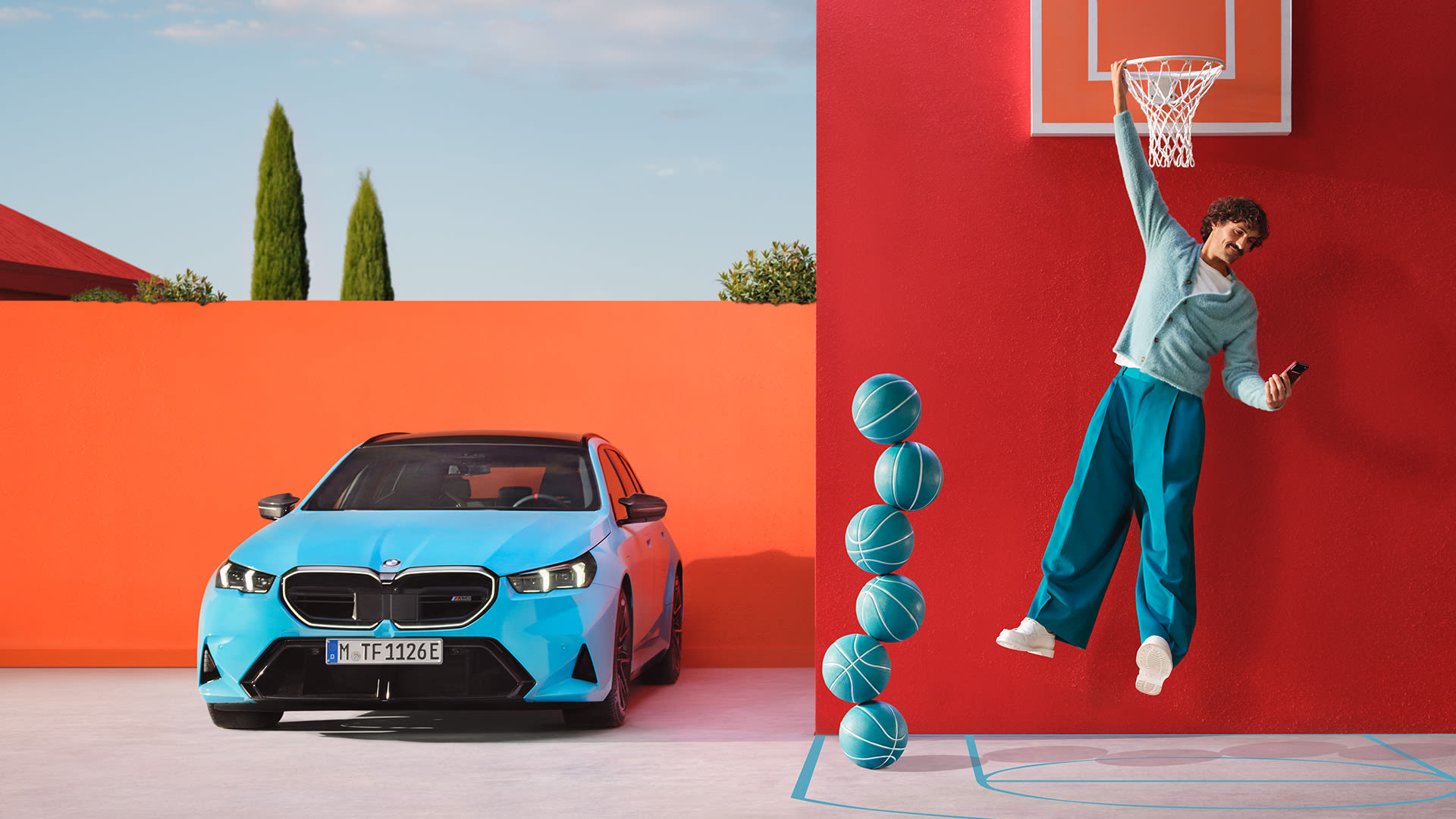 Blue BMW parked next to a red wall with a stand having 6 basketballs and a guy hanging from the basketball ring