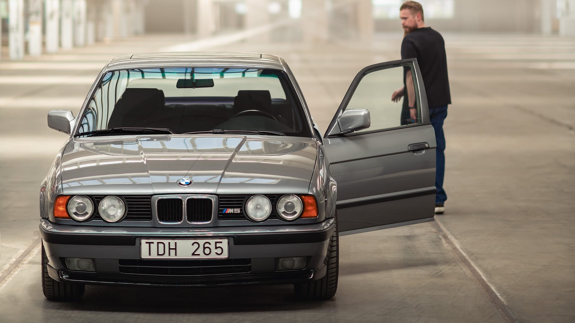 Front view of a geay BMW M5 with the left door open and a person looking over the sholder at the back of the car