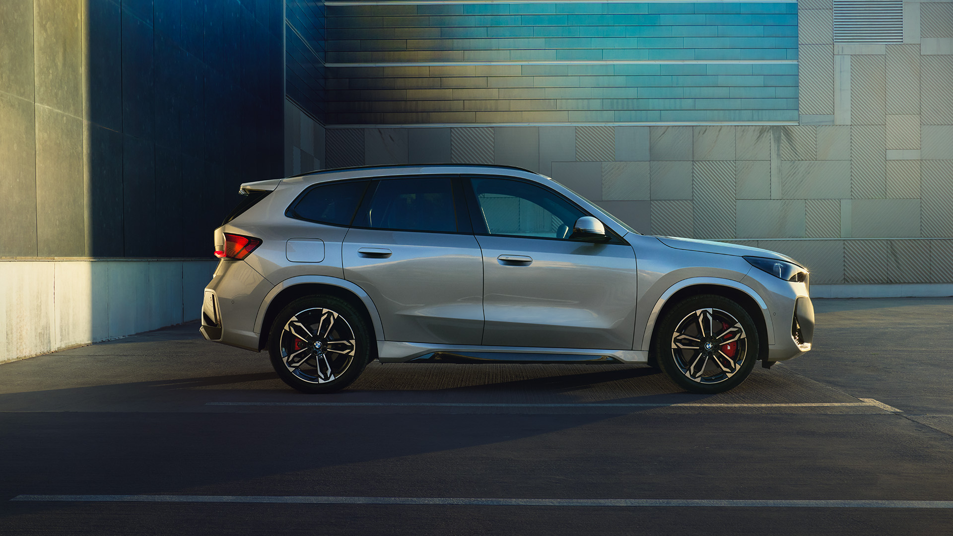 A white BMW SUV parked in an empty parking lot next to a modern building with a blue and beige facade during sunset.