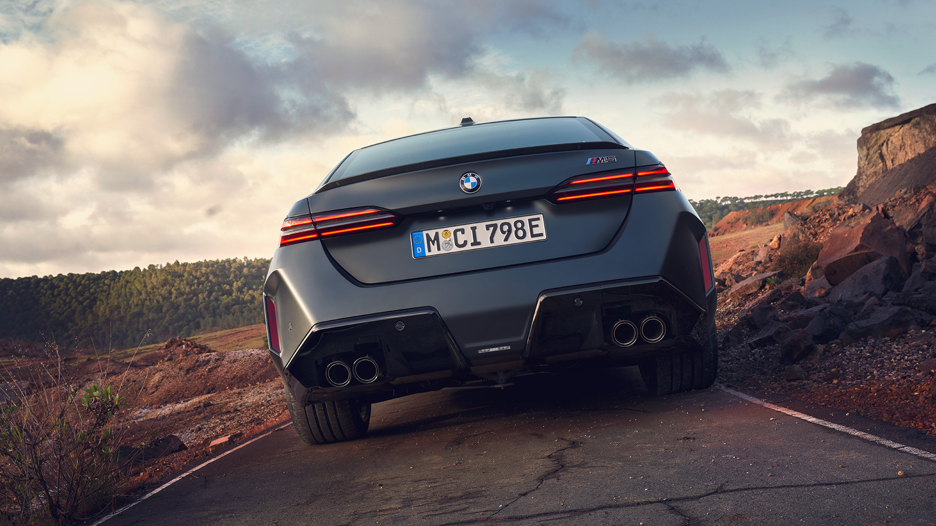 Rear view of a gray BMW car on a deserted road with rocky terrain and cloudy sky in the background.